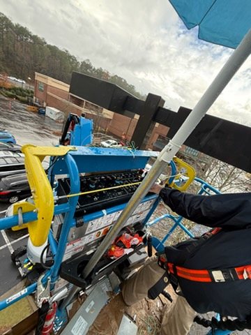 Worker wearing a fall-protection harness in an aerial-lift basket beside an electrical fixture at a commercial building.